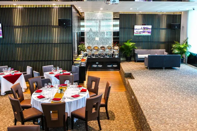 A restaurant dining room with round tables set with white linen and red napkins, a buffet station in the background and a lounge area with sofas on the right.