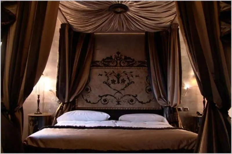 A bedroom with a canopy bed draped in dark gold curtains, an ornate patterned headboard, and wall sconces lit on either side.