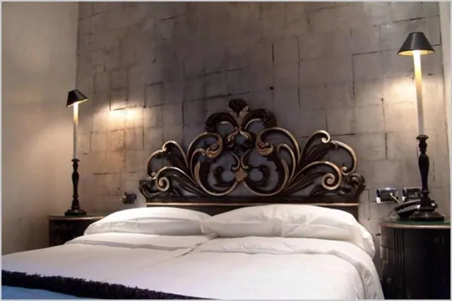 A double bed with white linens and an ornate carved dark headboard sits between two lit bedside lamps against a textured grey wall.
