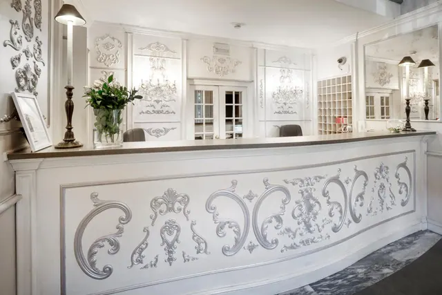 A white reception desk with silver ornamental scrollwork stands in a lobby with mirrors, a chandelier, a table lamp and a vase of white flowers.