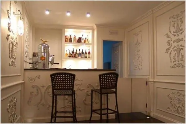 A small bar area with illuminated shelves of bottles, two high bar stools and ornate plasterwork on the walls.