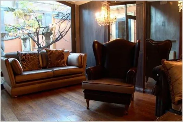 A seating area with a leather sofa, a wingback armchair with an ottoman, and a chandelier, with a window looking onto a courtyard tree.
