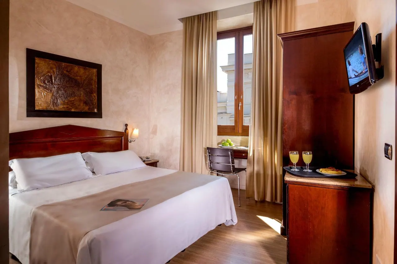 A double bed with white linen sits in a bedroom beside a wooden wardrobe, a wall-mounted television and a small desk by a window.