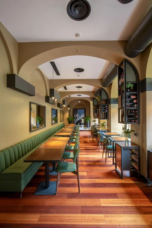 A long restaurant dining room with arched ceilings, green banquette seating along one wall, wooden tables with green chairs, and wine bottles displayed on wall shelves.