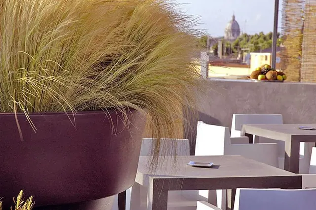 A rooftop terrace with a large grass planter, white chairs and grey tables, with a domed church and rooftops visible in the background.