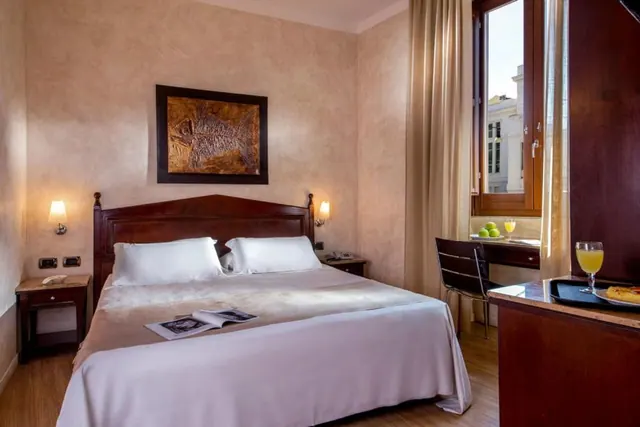 A double bed with white bedding and a framed artwork above the headboard, with a desk and chair by a window and a tray with orange juice at the right edge.