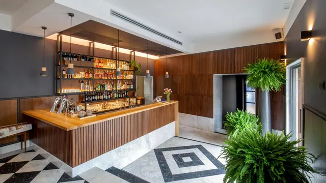 A wood-slatted bar counter stands in front of shelves of bottles, lit by pendant lamps, with potted ferns and a black and white marble floor nearby.