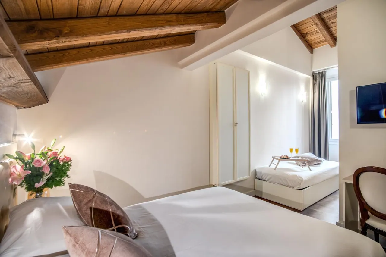 A bedroom with a wooden beamed ceiling, a double bed with pillows, a vase of pink flowers, a second single bed with a breakfast tray, a white wardrobe and a wall-mounted television.