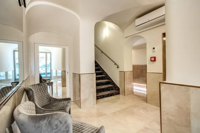 A lobby with grey upholstered armchairs, a staircase with dark carpeted steps, marble walls and floor, and a wall-mounted air conditioner above an arched doorway.