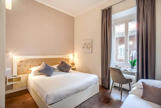 A double bed with a tufted headboard stands beside a desk and chair under a window that looks onto a neighbouring building.