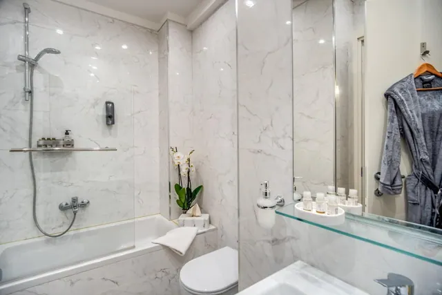 A bathroom with marble-look walls, a bathtub with a handheld shower and glass screen, a toilet, a white orchid, and a grey bathrobe hanging on the wall.