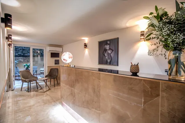 A lobby reception desk with a marble-look front sits beside a seating area with two armchairs and a side table, with glass doors opening onto a terrace with greenery.