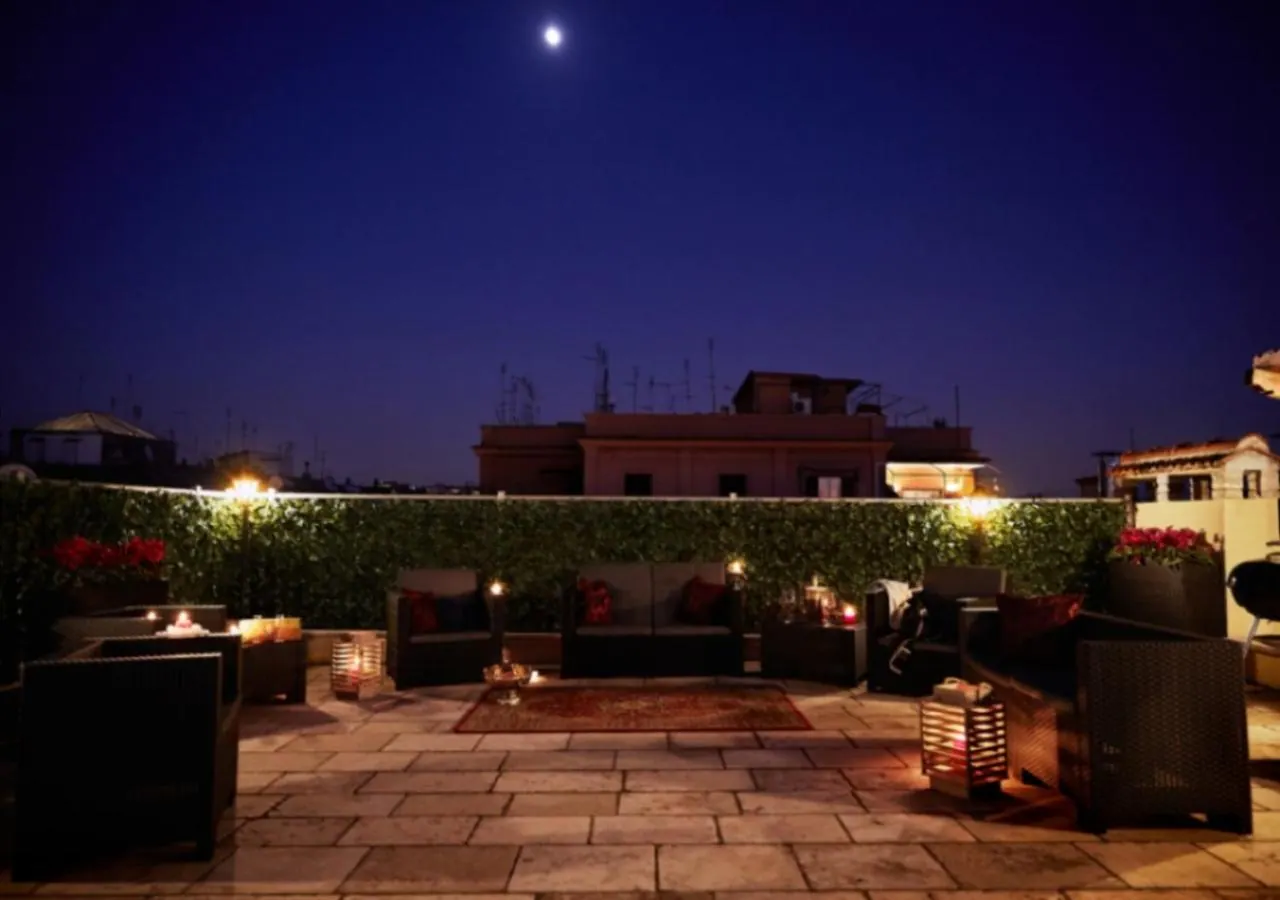 A rooftop terrace at night with wicker sofas, lit candle lanterns, a hedge wall, and rooftops under a sky with a moon.