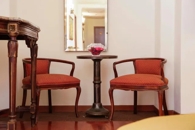 Two upholstered wooden armchairs flank a small round pedestal table holding a glass bowl of red petals, with a mirror on the wall above.