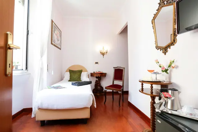 A small bedroom with a single bed, a wooden floor, a red velvet chair, a gilded wall mirror, a wall-mounted television, a kettle on a tray and a small fridge.