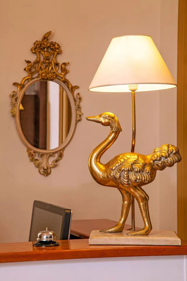 A brass ostrich-shaped table lamp with a cream shade stands beside a call bell and a tablet on a wooden reception desk, with an ornate gilt mirror on the wall behind.