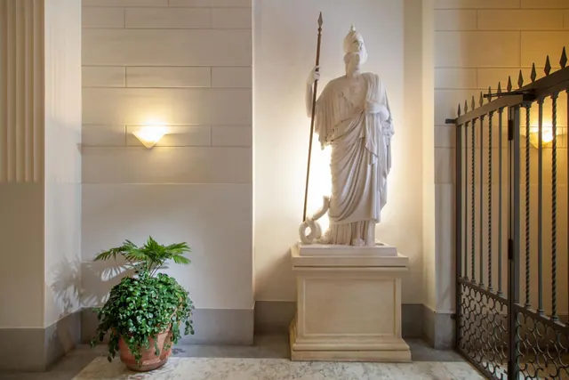 A white marble statue of a helmeted figure holding a spear stands on a pedestal beside a potted plant and a wrought iron gate.