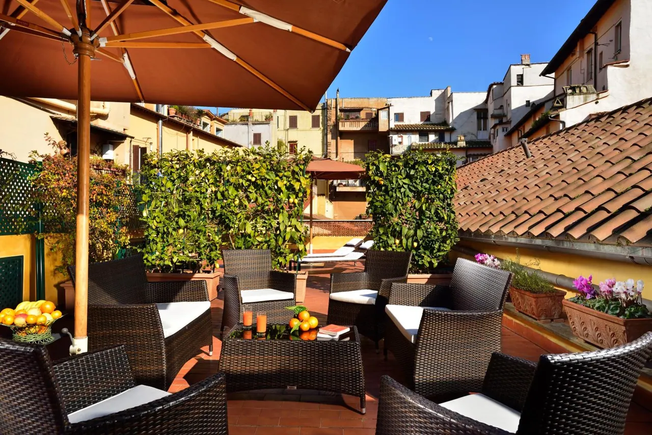A rooftop terrace with wicker armchairs and white cushions, a large brown parasol, potted plants and a terracotta tiled floor, with neighbouring rooftops and buildings beyond.