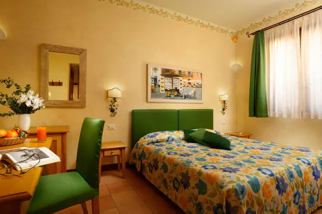 A bedroom with a double bed under a floral bedspread, a green headboard, a wooden desk with flowers and fruit, and a window with green curtains.