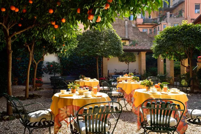 An outdoor courtyard dining area with yellow-clothed tables, wrought iron chairs with white cushions, and orange trees overhead.