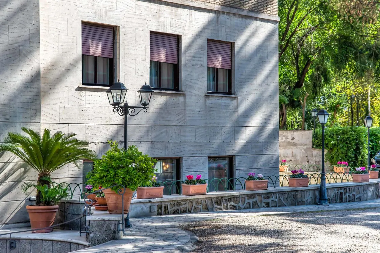 A pale stone facade of a building with shuttered windows, potted plants, and black lamp posts along a paved path with a low planted wall.