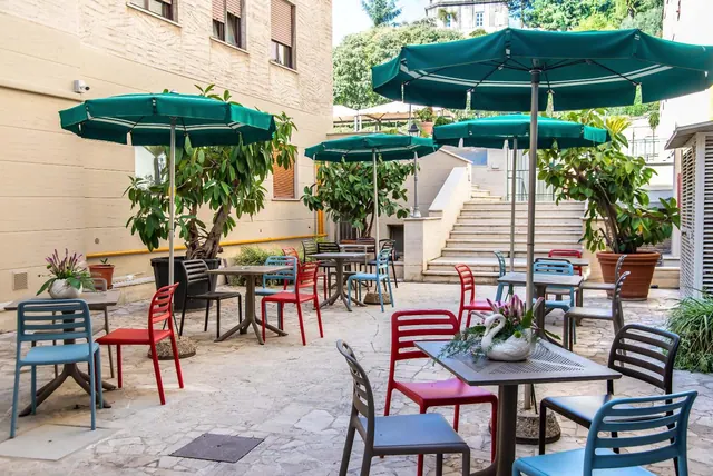An outdoor courtyard terrace with green parasols, red and blue chairs and tables on a stone-paved patio, with stairs and trees behind.