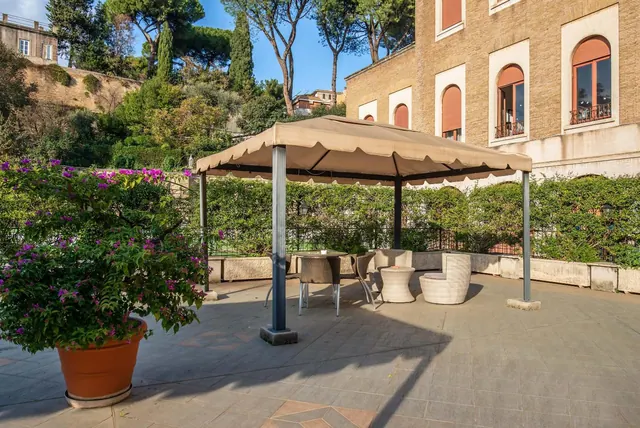 A paved terrace with a canopy, wicker chairs and a table, a potted bougainvillea in the foreground, and a brick building with arched windows and hedges behind.