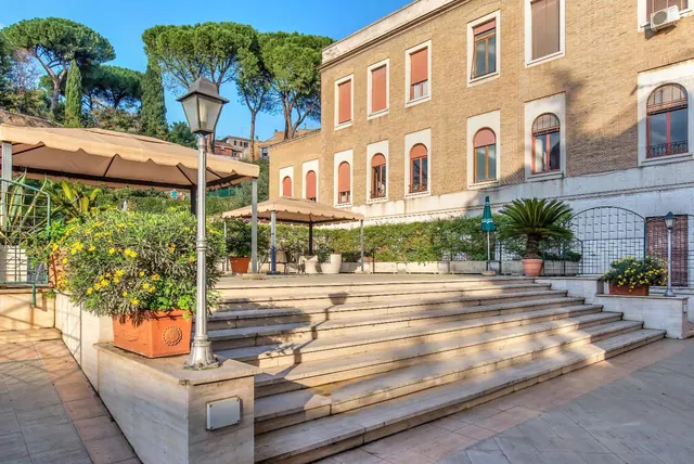 Wide stone steps lead up to a terrace in front of a brick building with arched windows, with potted plants, canopies and lamp posts.