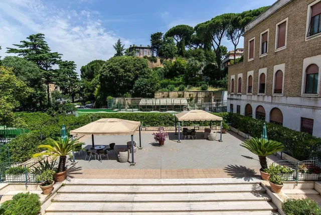 A paved terrace with canopy shelters, tables and chairs, potted palms and wide steps leading down, with trees, a hillside and a brick building beside it.