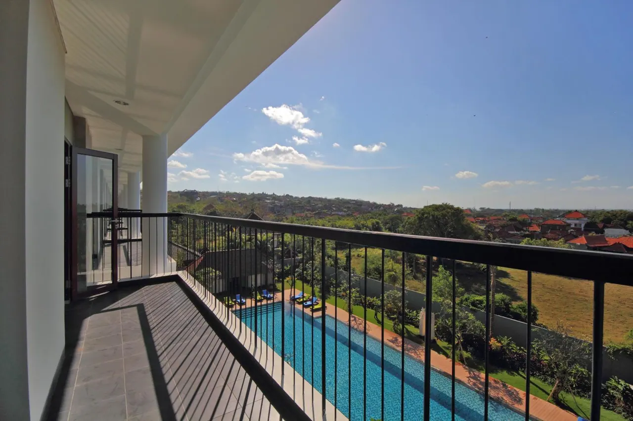 A balcony with a black metal railing overlooks a long swimming pool, green gardens and rooftops under a blue sky.