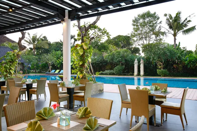 Outdoor restaurant with wicker chairs and tables set with green napkins under a pergola, with a swimming pool and garden behind.