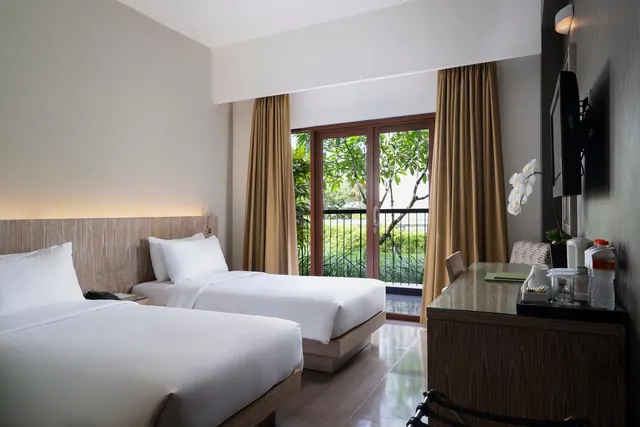 A bedroom with two made single beds, a wall-mounted television, a desk with a tray of glasses, and glass doors opening onto a balcony with trees beyond.