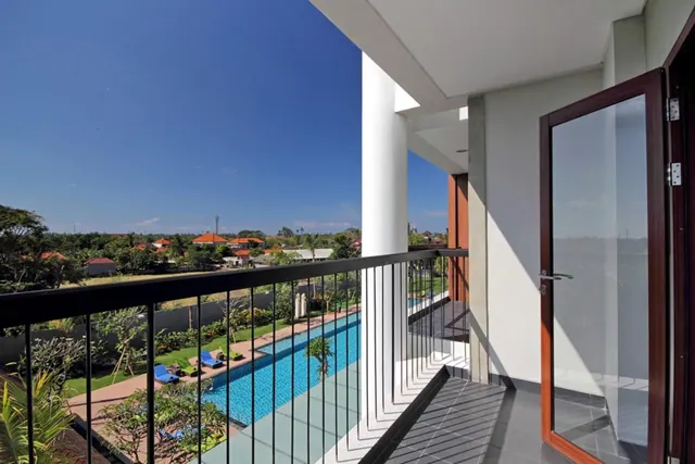 A balcony with a black metal railing overlooks a long lap pool, blue sun loungers, a landscaped garden and orange-roofed houses under a clear blue sky.