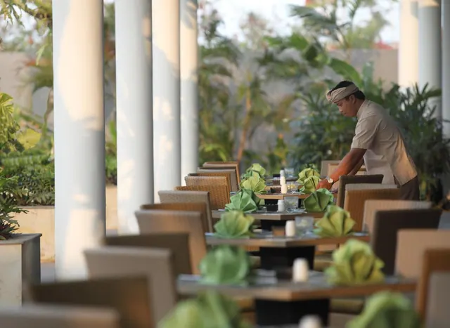 A staff member in a headwrap sets tables with green folded napkins on an open-air restaurant terrace lined with white columns and wicker-style chairs.