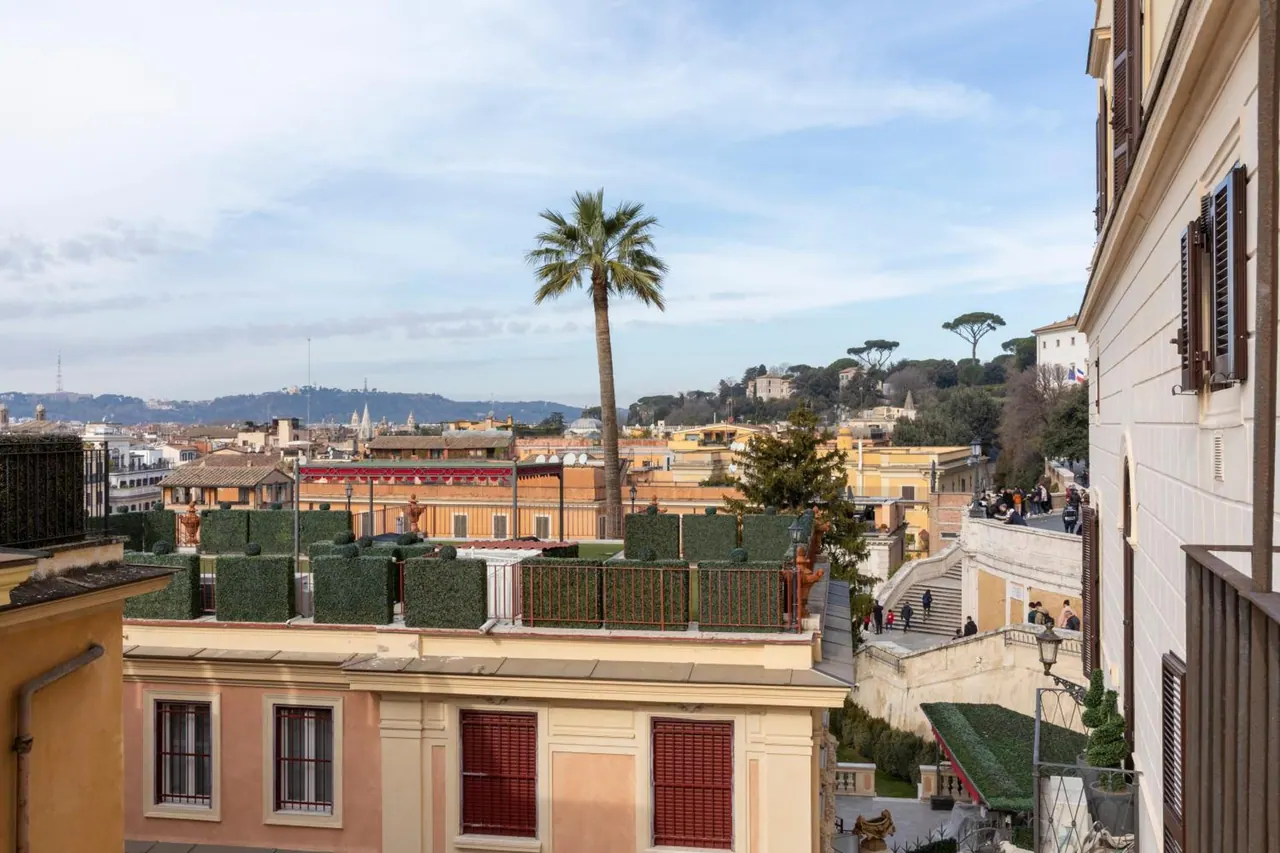 View from an elevated terrace over Rome rooftops, with a palm tree, clipped hedges, a staircase with people in the distance, and hills on the horizon.