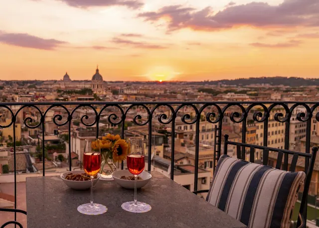 A table with two glasses of rosé, two small bowls of snacks and a vase of sunflowers sits on a balcony overlooking Rome at sunset, with the dome of St. Peter's Basilica visible in the distance.