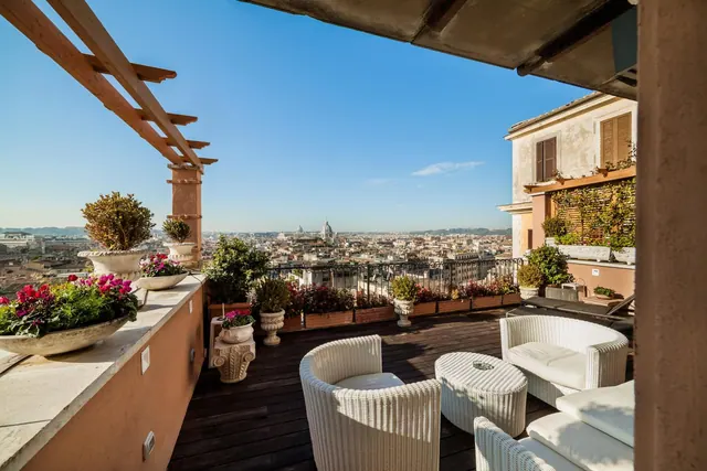 A furnished rooftop terrace with white wicker seating, potted plants and a wide view over Rome toward a distant dome.