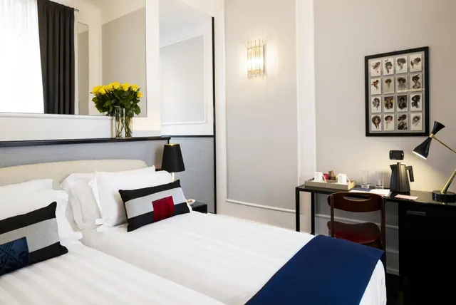 A hotel bedroom with two beds, a black desk with a chair and kettle, a framed wall print, and a vase of yellow roses.
