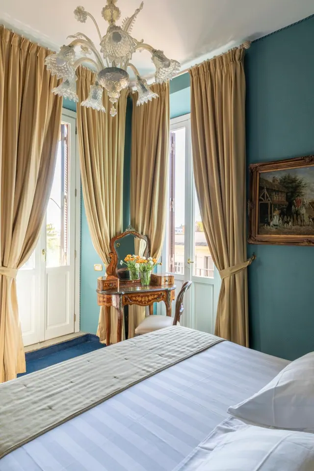 A bedroom with teal walls, gold curtains, a glass chandelier, a framed painting, a marquetry dressing table with a chair, and a striped bed in the foreground.