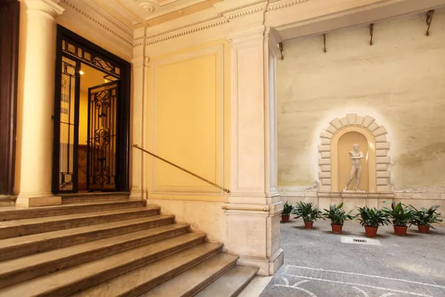 A stone staircase leads up to an ornate entrance with an iron gate, beside a courtyard with a statue in a wall niche and potted plants.