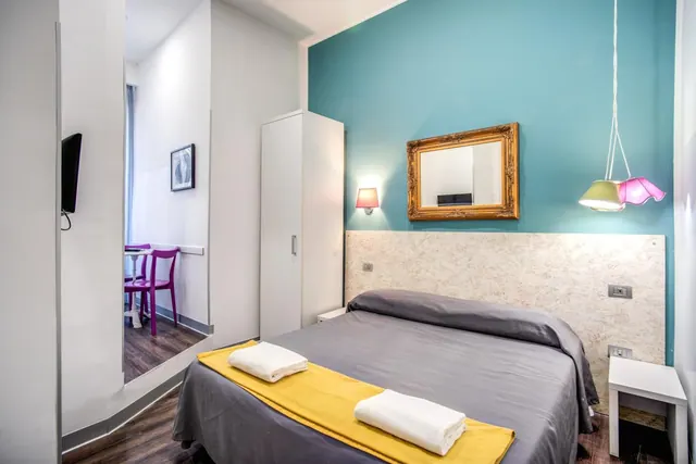 A double bed with grey bedding, a yellow runner and two white towels sits beneath a cream upholstered headboard and a teal accent wall with a gilt-framed mirror.