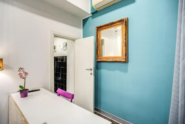 A small room with a teal wall and gilded mirror, an open white door to a black-tiled bathroom, and a white table holding an orchid and a remote, with a purple chair beside it.