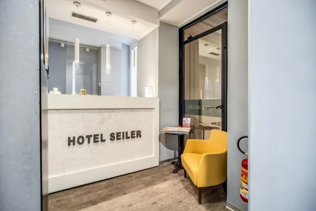 A white reception desk marked HOTEL SEILER stands beside a yellow armchair, a small table with a magazine and a red fire extinguisher on the wall.