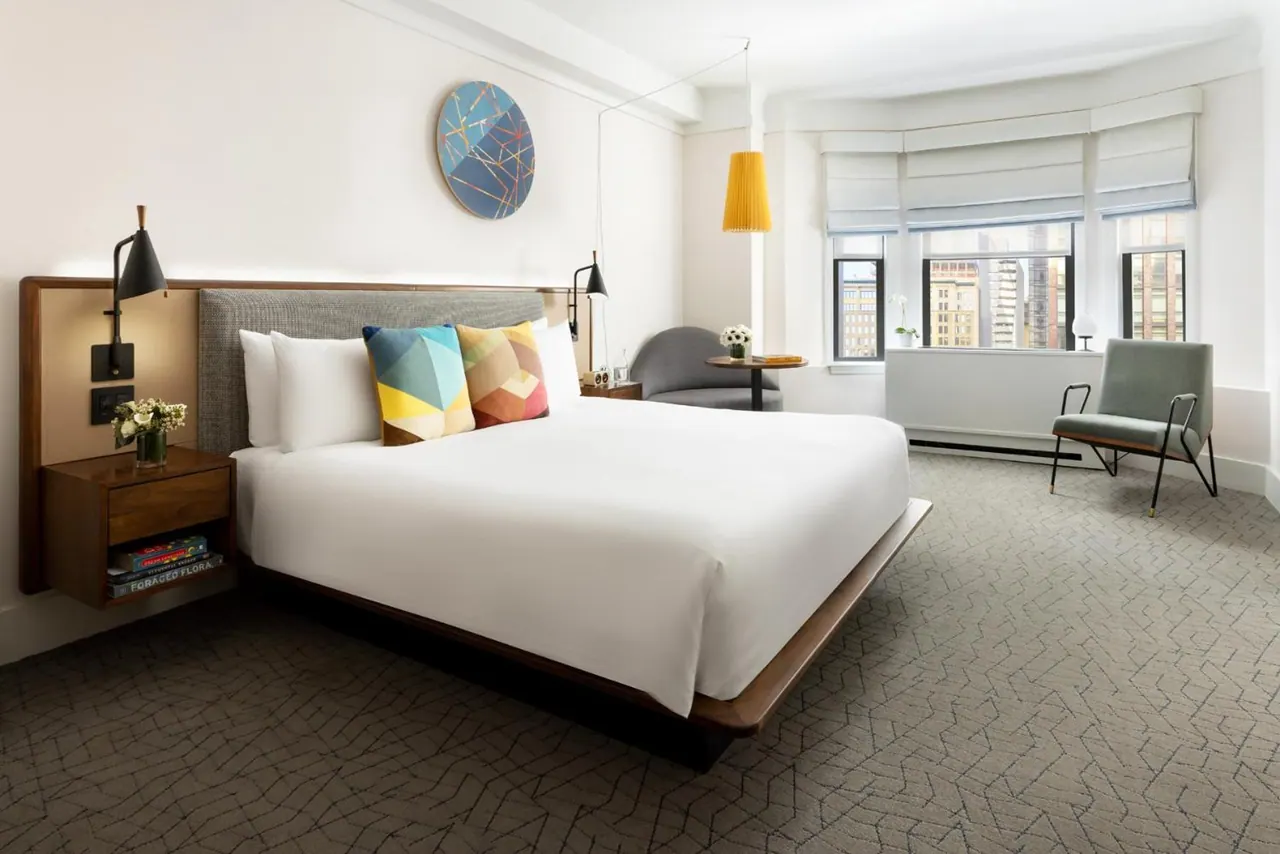 A king bed with white linens and colorful cushions sits in a bedroom beside a bay window with a view of city buildings.
