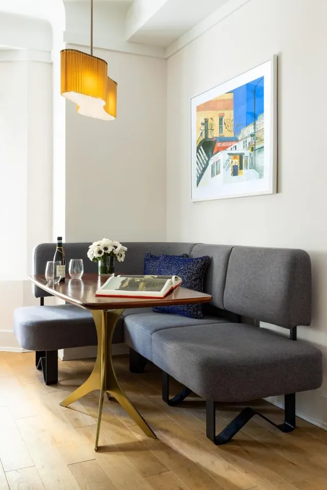A grey upholstered corner banquette sits beside a wooden table holding a wine bottle, two glasses, flowers and a book, under an amber pendant lamp and a framed street painting.