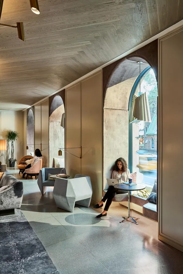 A woman sits working on a laptop at a small round table beside an arched window in a lobby lounge, with a street and building visible outside.