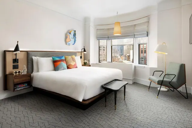 A bedroom with a king bed, wall-mounted reading lights, an armchair and a bench, with city buildings visible through the bay windows.