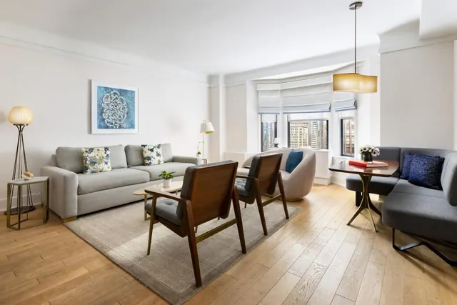 A staged living room with a grey sofa, wooden armchairs, a small dining table with a banquette, and bay windows showing city buildings outside.