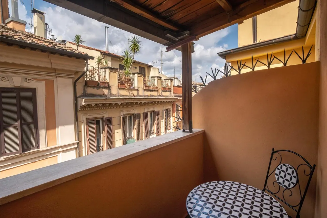 A private balcony with a mosaic-top bistro table and wrought iron chair overlooks neighbouring buildings with potted plants under a partly cloudy sky.