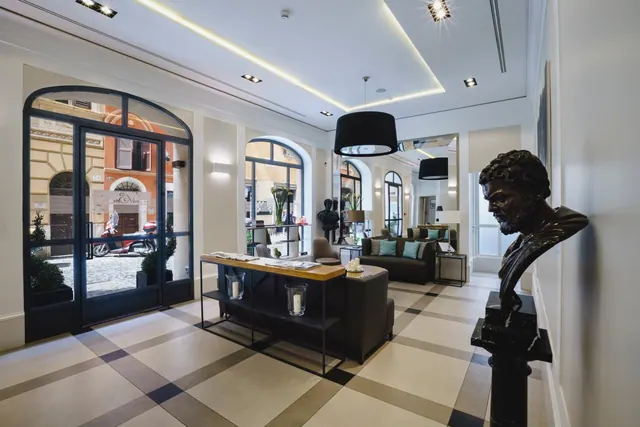 A hotel lobby with a checkered stone floor, a bronze bust on a black pedestal, seating, and arched glass doors opening onto a street with a parked scooter.